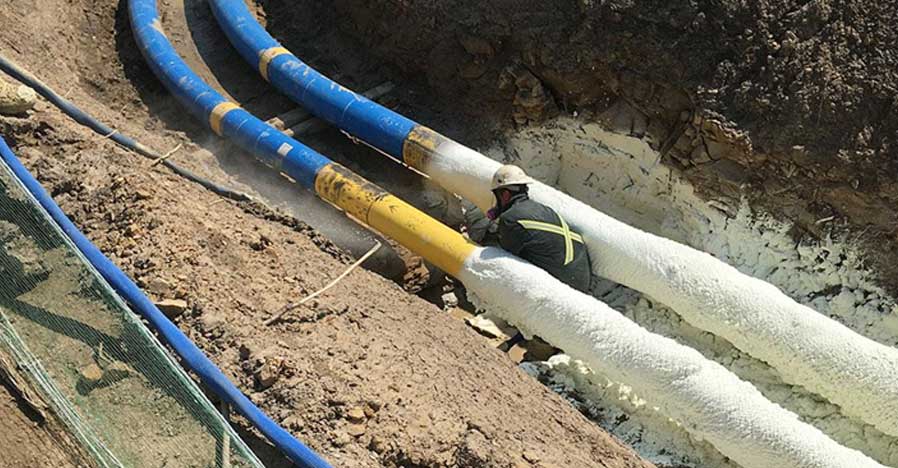 Worker insulating large blue and yellow pipelines with a white insulation material in an outdoor setting.